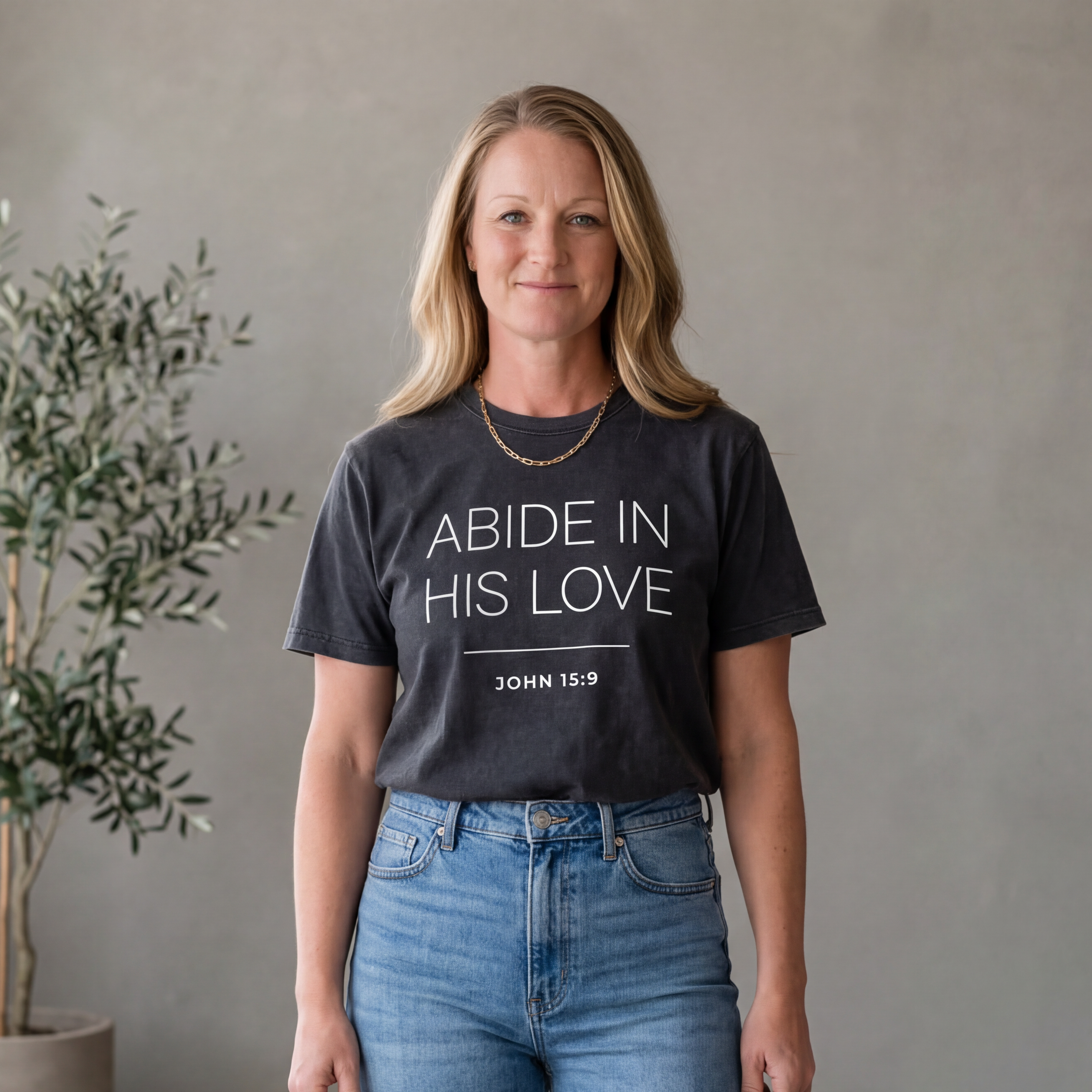 Founder wearing "abide in his love" mineral wash tshirt with text, standing in a room with plants and a neutral wall.