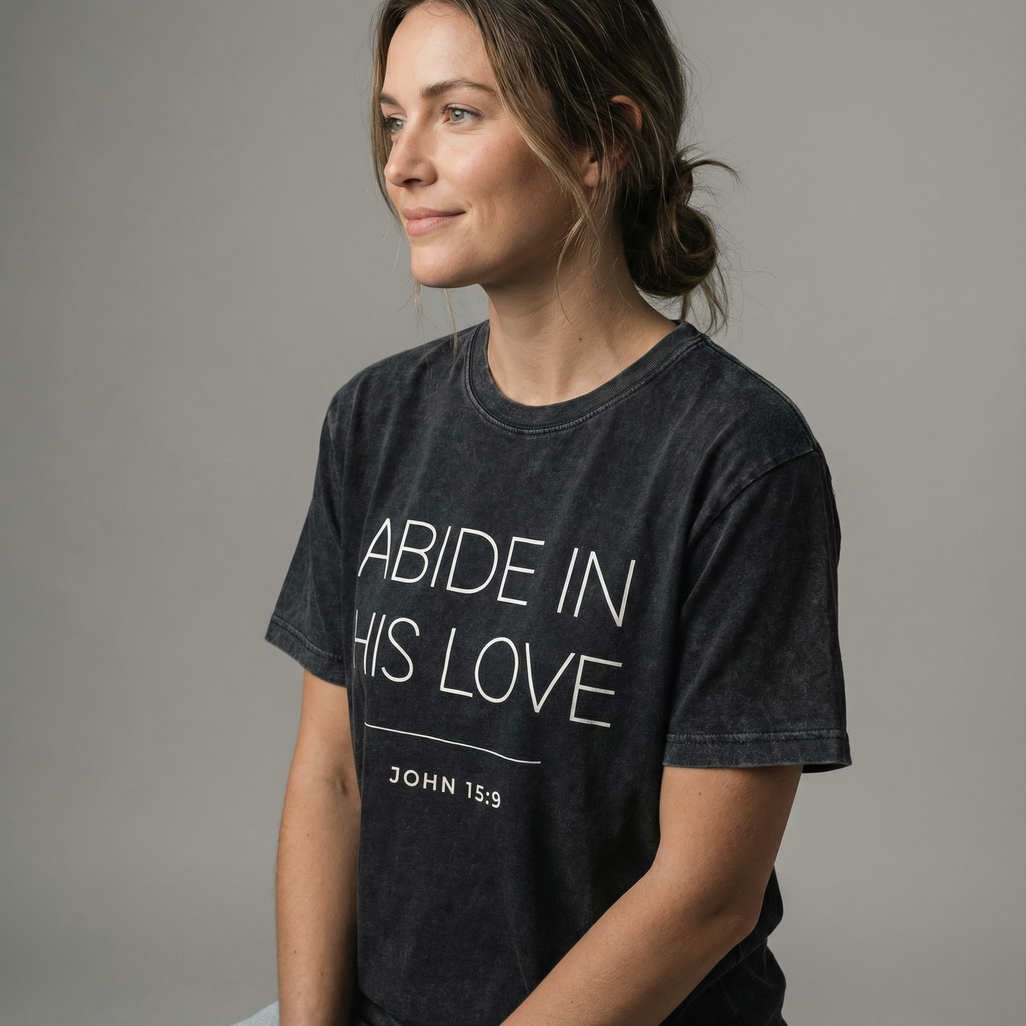 Side angle studio view of a woman wearing the "abide in his love john 15:9" minimalist graphic tee with a calm, natural expression.